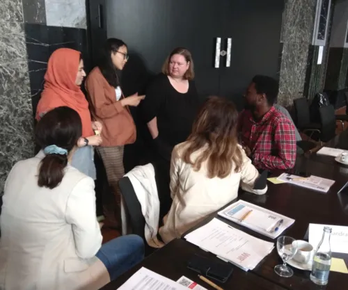 Fünf Frauen und ein Mann in einem lockeren Kreis. Drei stehen, drei Sitzen an einem Tisch. Sie sind einander zugewandt und scheinen miteinander zu reden.
